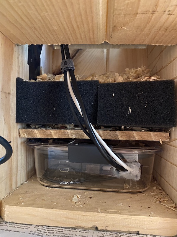 A side view inside the birdhouse through an open panel. Inside is the Raspberry Pi 3B+ within its waterproof/shatterproof case, with a hinged piece of wood on top, followed by about a cubic foot of space for birds to nest, along with sawdust as nesting material.